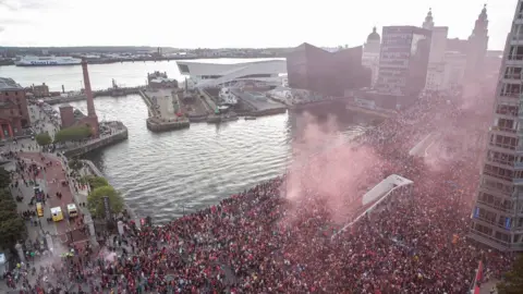 Liverpool City Council Thousands of people line the parade route on the The Strand, with red smoke visible above the docks.
