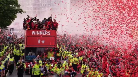 PA Media A red open-top double decker bus takes the Liverpool FC squad through the streets of the city for a victory parade. The sky is filled with red ticker tape and there are huge crowds of people surrounding the bus.