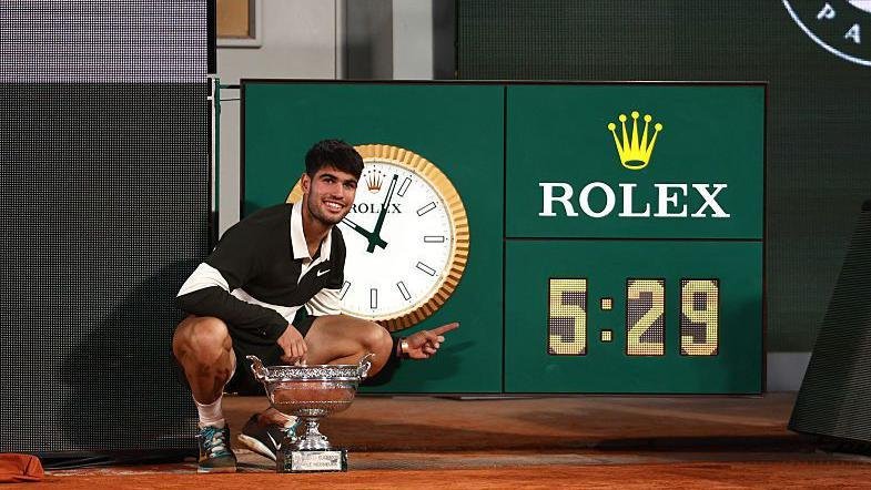 Carlos Alcaraz poses next to the clock on Court Philippe Chatrier after his five-hour and 29-minute final against Jannik Sinner